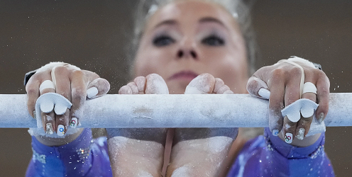 a female-appearing gymnast on horizontal bar, with both hands and feet used in the midst of a performance