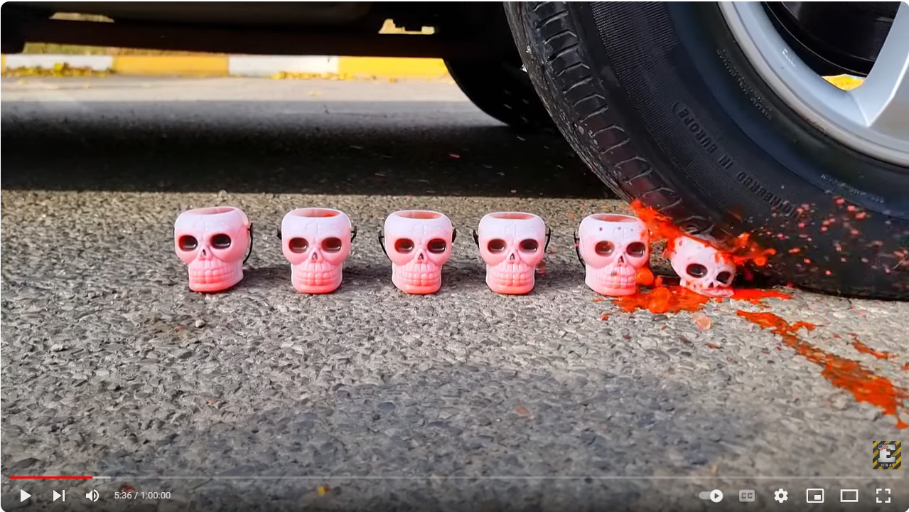 Screenshot of a video of a car tire slowly crushing rubber toy skulls containing some sort of orange or red gel.