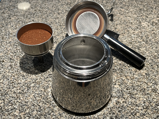 A stovetop coffee maker filled with boiling water, with a basket of coffee grounds beside it