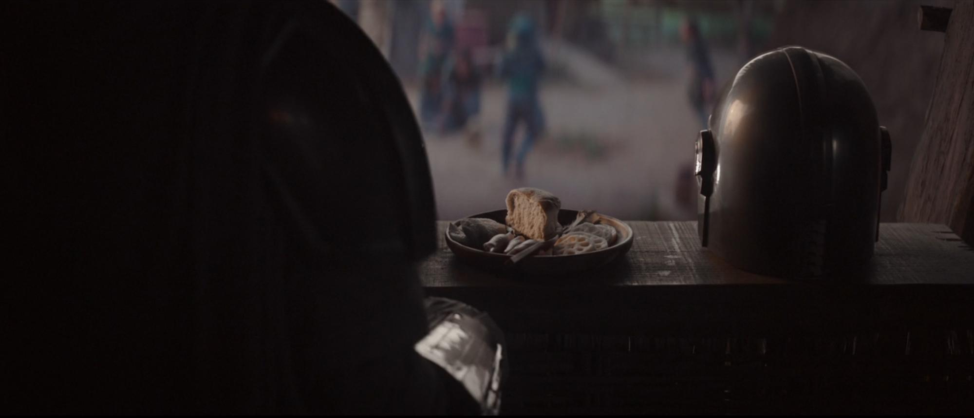 Din Djarin sitting down to eat a meal and watch the children play with his helmet off.