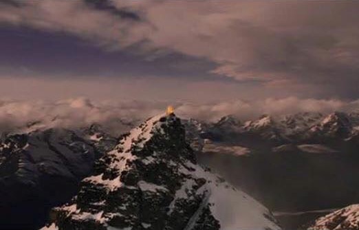 A snowy, rocky point of a mountain peak, amid other similar peaks in the distance, high enough to be almost in the clouds.  A fire burns atop it.