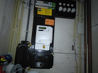 a utility closet, with a black electrical panel containing the breakers, electrical conduit to its left and right, with some other wires and boxes of an unknown nature as well