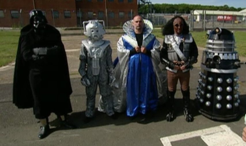 Photo of 5 cosplayers on tarmac at a fenced-off facility of some sort.  From left to right are Darth Vader; a silver suit with a silver mask with a stylized frowning mouth, round eyes and bulky ear covers connected over the top of the head like headphones; a bald man in a blue robe with a white cape and a head-high, stiff, circular flaring collar; a Next-Generation-era Klingon; and a black pepper pot with silver studs and plungers sticking out in place of arms.