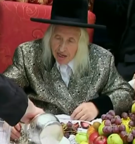 An elderly man, wearing a gold-and-black-colored Chassidic-style jacket and a flat-brimmed black hat, clean-shaven, with white hair falling below his chin on either side of his face