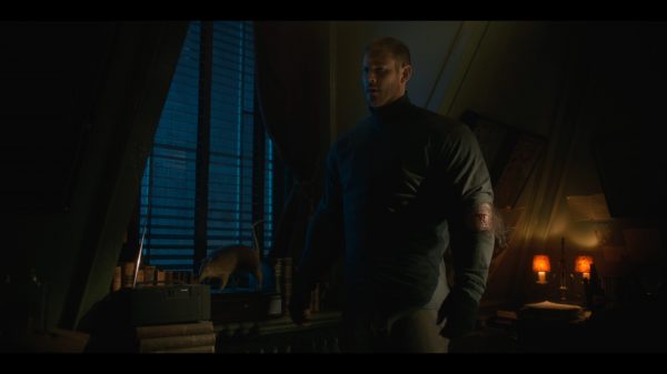 Luther stands in a dark room, his top is ripped on the left arm near the elbow revealing a cut