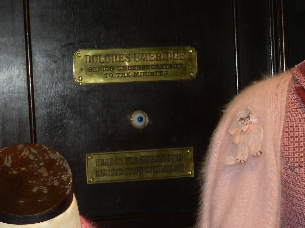 The door to Umbridge's office. A plaque says "DOLORES UMBRIDGE: Senior Undersecretary to the Minister". Underneath the plaque is a blue eye set into the door. Another plaque underneath the eye says "HEAD OF THE MUDDLE-BORN REGISTRATION COMMISSION".