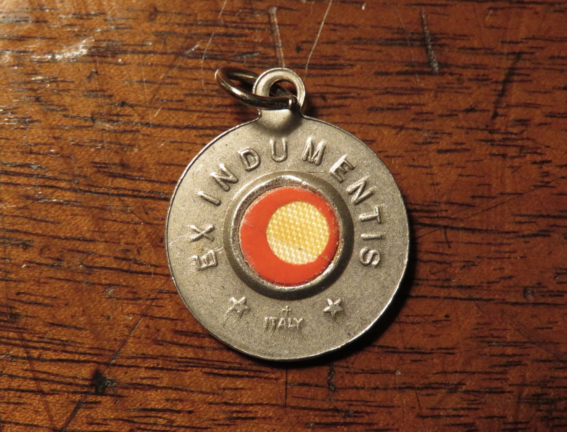 A round necklace medal with a yellowed piece of cloth in the center, with a red protective seal and a metal outer circle with the phrase "Ex indumentis" embossed on it