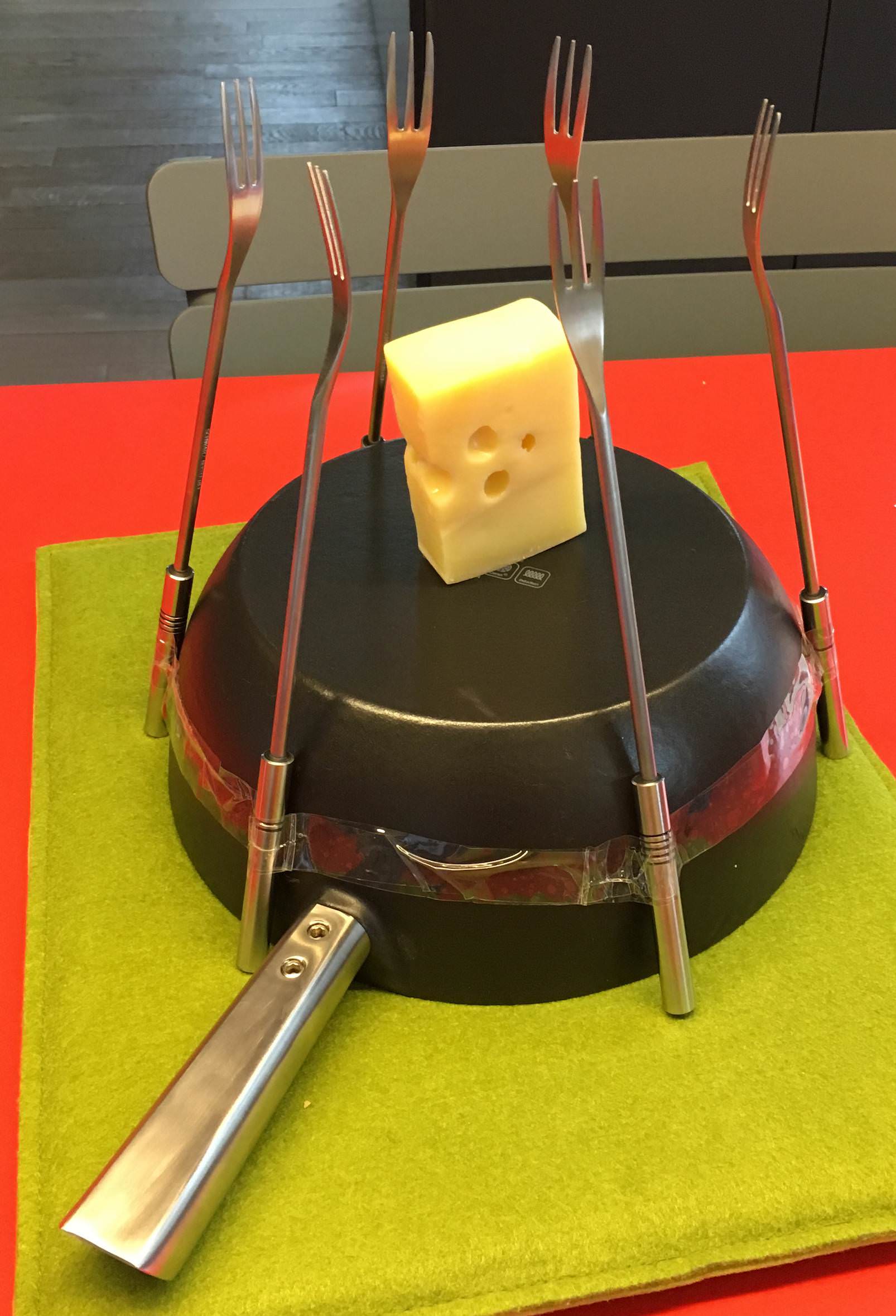 Fondue pot with 6 forks taped to the outside and a slice of Emmental cheese on top.