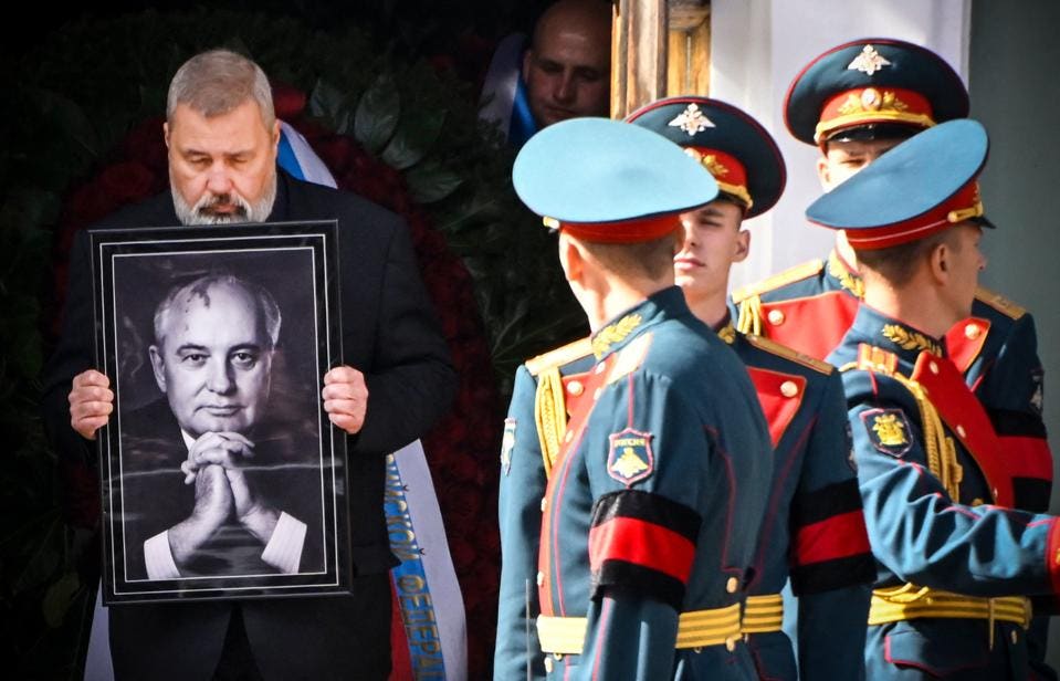 A Russian man holds a black and white photo of Gorbachev while four military men with black and red armbands stand nearby