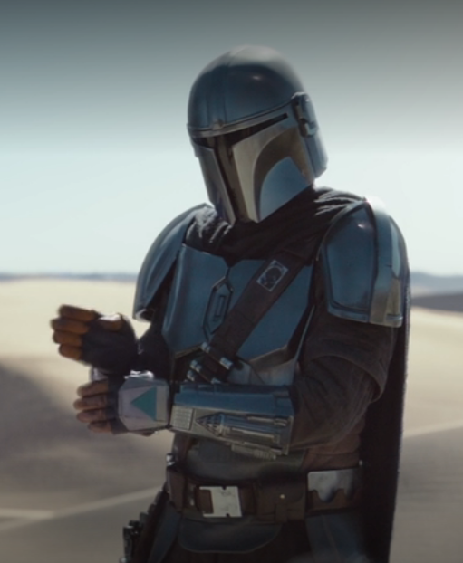 The Mandalorian stands facing a point to the viewer's left.  His left arm is in front of him, horizontally from the elbow, with the fingers straight and hand oriented vertically with the back facing outward.  His right hand, also with straight fingers and held vertically, is above his left hand as though making a chopping motion.