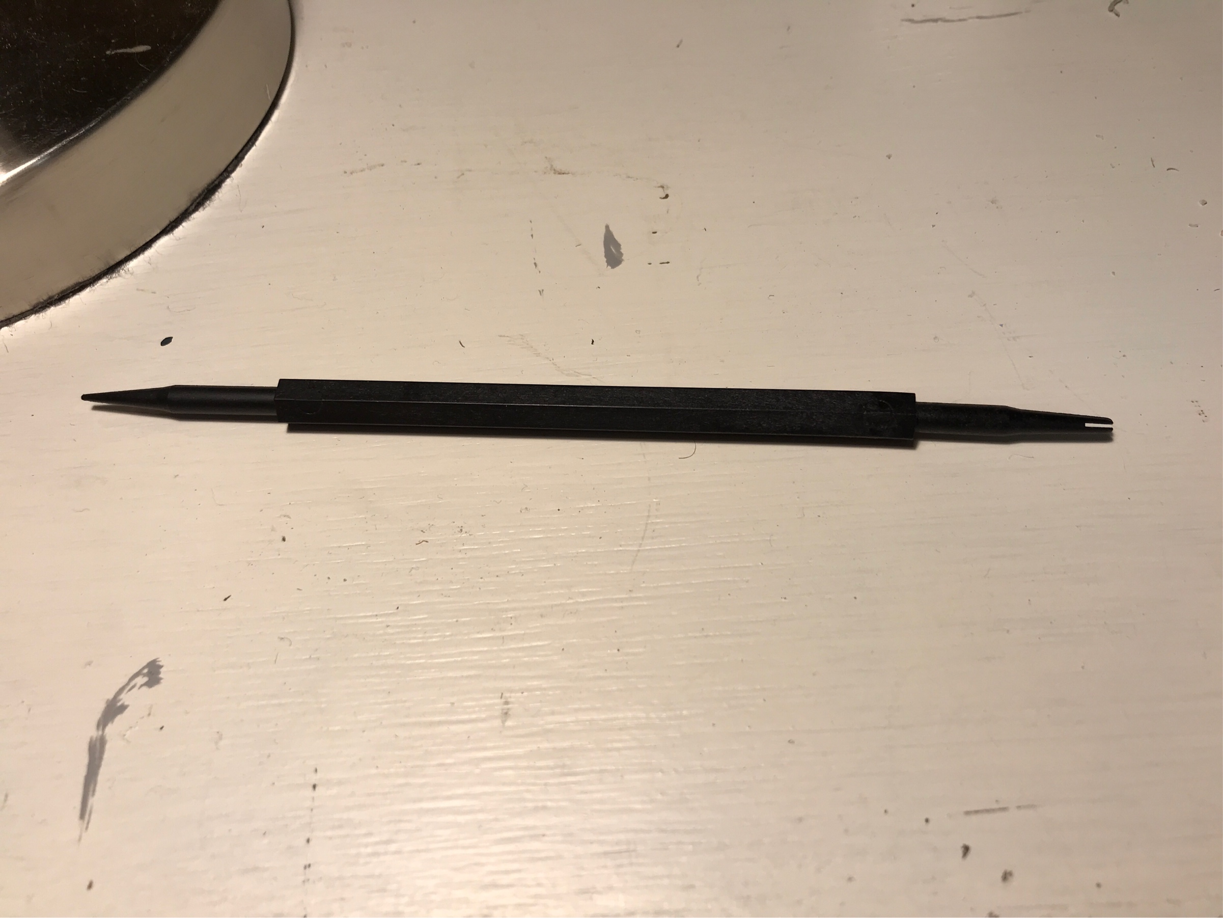 A picture of the mystery tool, lying on a white-painted wood surface. The tool is made from black plastic, and consists of a thin hexagonal shaft with round shafts extending from either end.